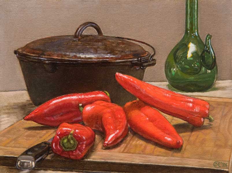 Still life with five long red Anaheim chili peppers, on a cutting board, with a kitchen knife, a cast iron Dutch oven and a green glass wine carafe in the background.
